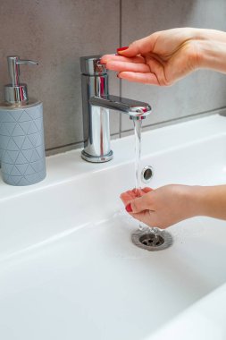 Banyoda gümüş musluğu olan beyaz lavabo. Ellerinde sıvı sabun olan gri teneke kutu. Musluk suyunu açmak, kişisel el hijyeni. Akan suyun altında el yıkama