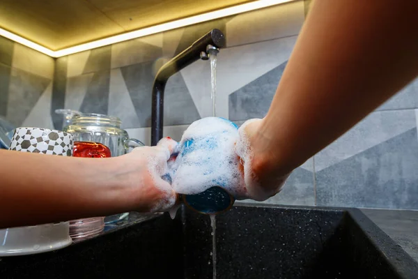 Hands with sponge wash the cup under water, housewife woman in washing blue mug in a kitchen sink with a blue sponge, Hand cleaning, manually, housework dishwasher