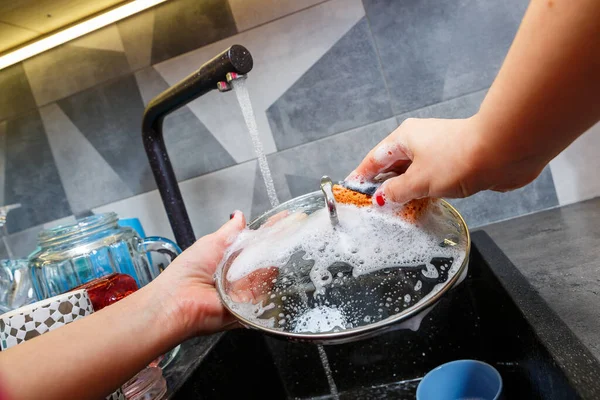 Hands with a sponge wash the pan cover under water, the housewife mistress washes a blue mug in the kitchen sink with a blue sponge, manual cleaning, manually, a home dishwasher