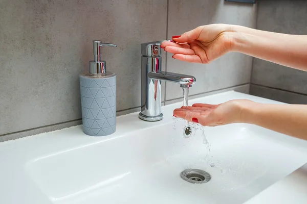 White sink with a silver faucet in the bathroom. Gray can with liquid soap for hands. Turning on the tap water, personal hand hygiene. Hand washing under running water