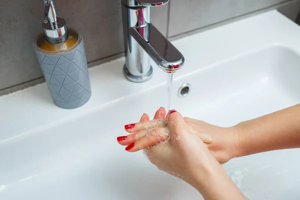 White sink with a silver faucet in the bathroom. Gray can with liquid soap for hands. Turning on the tap water, personal hand hygiene. Hand washing under running water