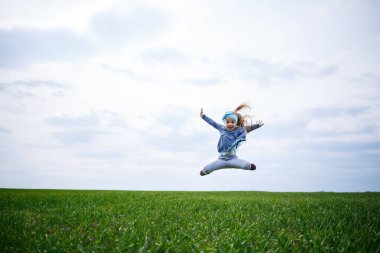 Küçük kız koşar ve zıplar, tarladaki yeşil çimenler, güneşli bahar havası, çocuğun gülümsemesi ve neşesi, bulutlu mavi gökyüzü