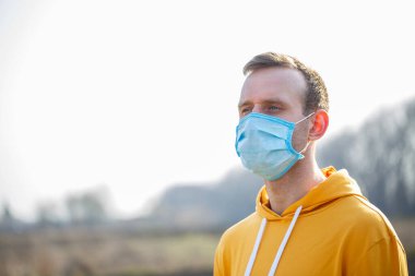 Açık havada cerrahi maskeli, lastik kulaklıklı bir adamın portresi. Ağız ve burnu örtmek için tipik üç katmanlı cerrahi maske. Bakteri maskesi prosedürü. Koruma kavramı.
