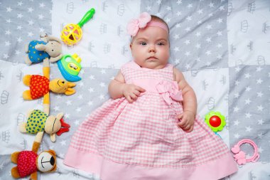 3 aylık bir bebek yatakta uzanıyor. Pembe elbiseli bir bebek ve bebek bezli bir saç bandı.