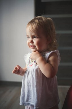1,5 yaşında küçük bir kız ayakta ve gülümsüyor. Çocuk dans eder ve yürür. Evdeki neşeli kız.