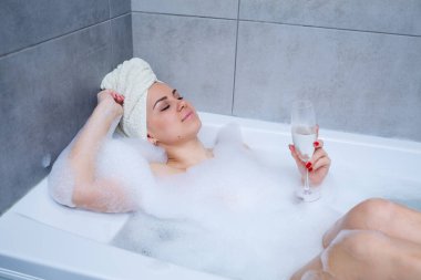 Kafasında beyaz havlu olan bir kadın beyaz bir küvette bir bardak şampanyayla yatıyor. Elinde bir sürü sidik sabunu var. Tam kıvrımlı köpük. Zor bir günün ardından rahatla. Spa gevşetici işlem