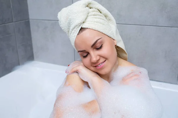 头戴白毛巾的女青年躺在浴室的白色浴缸里 泡沫的周围 辛苦一天后放松一下 温泉放松治疗 — 图库照片