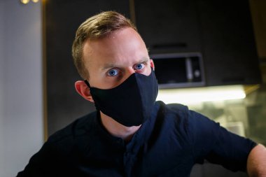 Siyah anti-virüs koruyucu maskeli genç bir işadamı, evde çalışıyor, mutfak masasında dizüstü bilgisayarla oturuyor. Evde, karantinada çalışmak