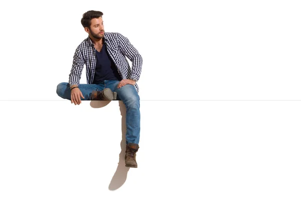Handsome man in boots, jeans and unbuttoned lumberjack shirt is sitting relaxed on a top and looking at the side. Full length studio shot isolated on white.