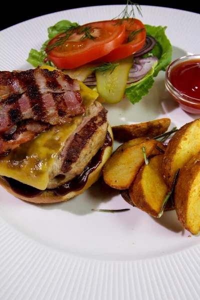 Kızarmış pastırmalı hamburger, patates kızartması, sebze ve ketçaplı beyaz tabakta. Yakın plan. Sağlıksız yemek..