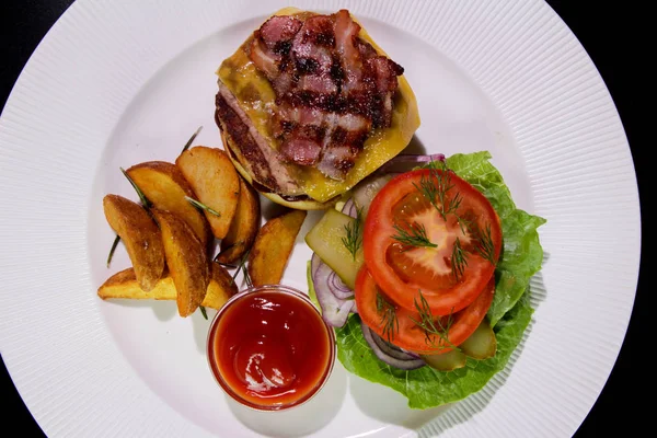 Kızarmış domuz pastırmalı hamburger, patates kızartması, sebze ve ketçaplı beyaz bir tabakta. Yukarıdan bak. Yakın plan. Sağlıksız yemek..