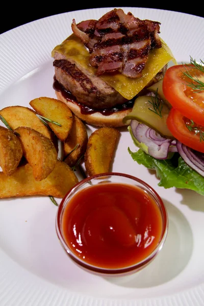 Kızarmış domuz pastırmalı hamburger, patates kızartması, sebze ve ketçaplı beyaz tabakta. Yakın plan. Enerji yoğunluğunda yiyecek..