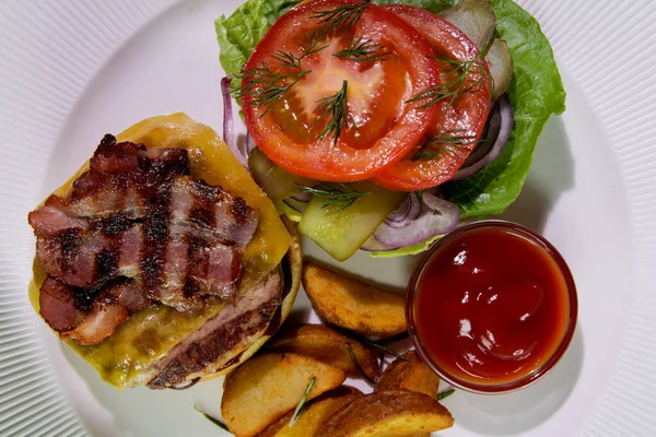 Kızarmış domuz pastırmalı hamburger, patates kızartması, sebze ve ketçaplı beyaz bir tabakta. Yukarıdan bak. Yakın plan. Enerji yoğunluğunda yiyecek..