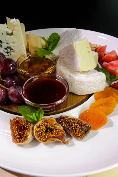 Cheese plate. Camembert cheese, Parmesan, blue cheese served with grapes, mint, jam, figs, honey, strawberries, dried apricots on a white plate.