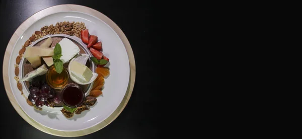 Peynir tabağı. Siyah bir arkaplanda izole edilmiş. Camembert peyniri, parmesan peyniri, üzümlü mavi peynir, reçel, incir, bal, çilek, kurutulmuş kayısı ve fındık. Yukarıdan görüş..