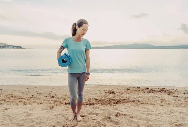 若い女性の海洋の背景にヨガマットと砂浜の上を歩いて — ストック写真