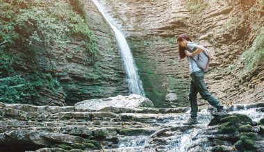 Sırt çantalı kadın, şelalenin önündeki dağ deresini geçiyor..