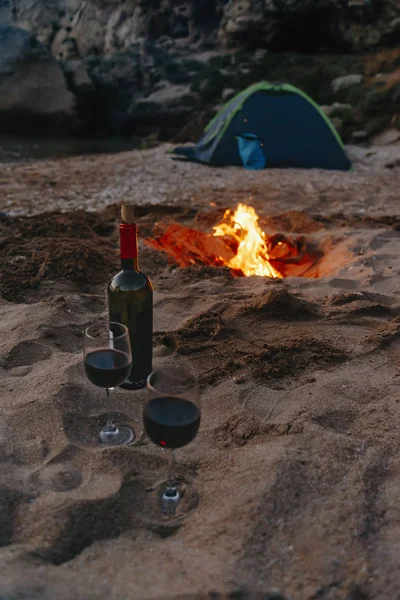 Bottle of red wine and two glasses on beach near campfire.
