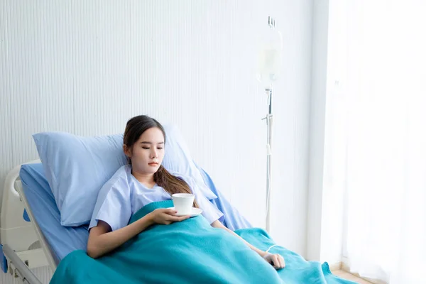 Asiática jovem paciente na cama com a mão segurando um café xícara — Fotografia de Stock