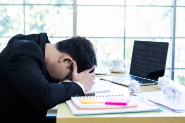 Stresli iş adamı dizüstü bilgisayarla çalışıyor ve ofis odasındaki kayıplardan sonra baş ağrısı çekiyor..