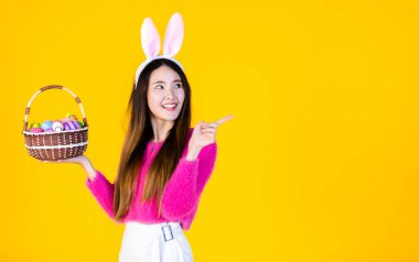 Paskalya bayramı konsepti, Mutlu Asyalı genç kadın tavşan kulaklı, elinde renkli Paskalya yumurtaları olan bir sepetle sarı boş fotokopi stüdyosu arka planında izole edilmiş..