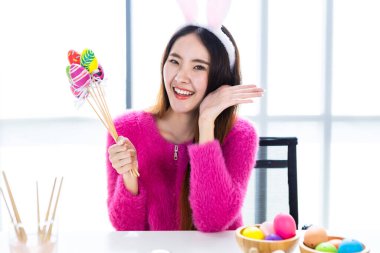 Paskalya tatili konsepti, Asyalı mutlu bir gülümseme tavşan kulağı takan genç bir kadın elinde renkli Paskalya yumurtaları beyaz ahşap masa arkasında şiş şiş.