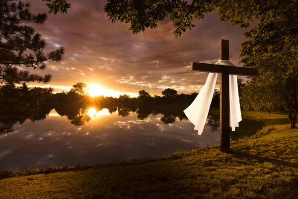 Beautiful photo illustration of an Easter morning sunrise on a cross by a calm lake. The warm light almost announces Jesus's rising from the dead. The fabric of Jesus's burial garments moves calmly in the breeze. 