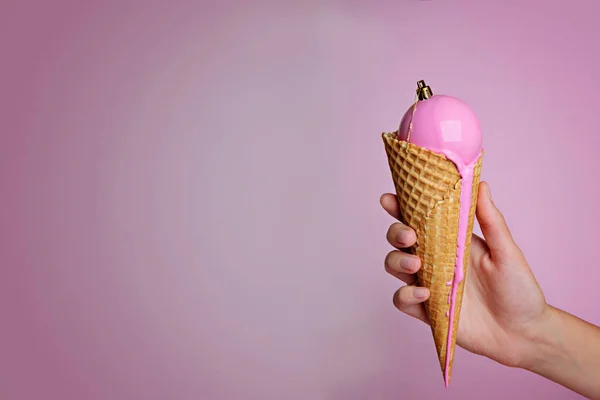 Female hand holding ice cream waffle cone with christmas ball in it on pink background.