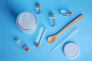 Küçük şişelerde hindistan cevizi yağı, krema kavanozu, kaşık ve damlatıcı mavi arka planda..