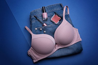 Pastel pembe sütyen, klasik mavi erkek arkadaş kotu ve mavi arka planda kadın aksesuarları ve kozmetik ürünleri..