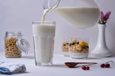 Bir adam kahvaltıda bardağa süt döker. Arka planda sunulan bir bardakta yoğurt ve taze böğürtlenli müsli. Beyaz ahşap masada kahvaltı..