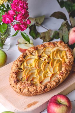 Tahtada ev yapımı elmalı turta. Beyaz ahşap bir masada, elmalar ve çiçekler etrafa saçılmış..