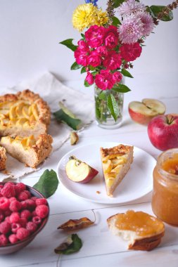 Çamaşır havlusunda dilimlenmiş bir elmalı turta. Beyaz ahşap bir masada tatlı, etrafında elmalar, tabakta ahududular, elma reçeli ve çiçekler var..