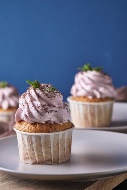 Tabağında pembe krema olan şenlikli kekler. Tatlı nane yaprakları ve çikolata parçacıklarıyla süslenmiş. Arkaplan rengi klasik mavi.