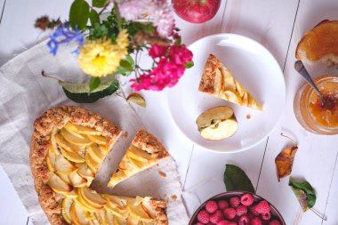 Çamaşır havlusunda dilimlenmiş bir elmalı turta. Beyaz ahşap bir masada tatlı, etrafında elmalar, tabakta ahududular, elma reçeli ve çiçekler var. Üst görünüm.