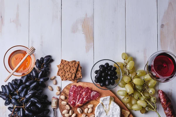 Romantik bir akşam yemeği için lezzetli ürünler. Peynir ve sosis lezzetleri, fındık, meyve, zeytin, ekmek, üzüm, şarap ve bal eski beyaz ahşap bir masada. Yukarıdan görüntüle.