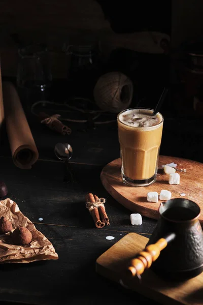 Coffee drink with ice and cream in the kitchen. Kitchen accessories around. Iced coffee in a glass, sweets, sugar, and cinnamon on a wooden table.