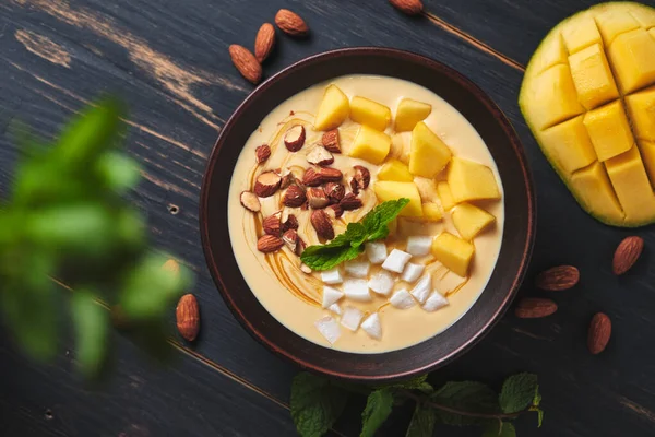 Tropical smoothie bowl with banana, mango, coconut and nuts. Refreshing dessert with mint on an old wooden table
