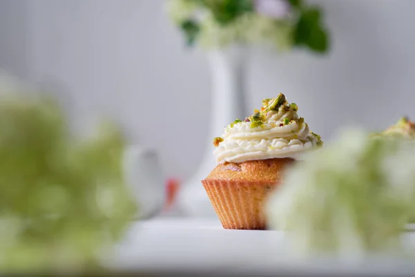 Fruit cupcakes with cream, decorated with ground pistachio and flowers in a vase on a white wooden table. Morning, rustic still life.
