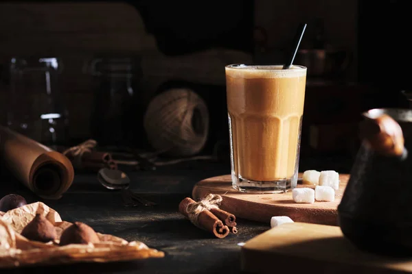 Coffee drink with ice and cream in the kitchen. Kitchen accessories around. Iced coffee in a glass, sweets, sugar, and cinnamon on a wooden table.