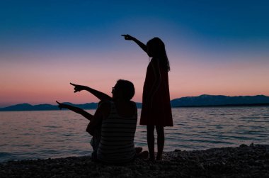 Gün batımından sonra kumsalda iki kızı olan bir annenin silueti, hepsi gökyüzünü işaret ediyor..