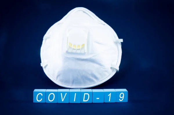 Face mask on dark blue background with covid-19 written on light blue blocks in conceptual image of health threatening virus.
