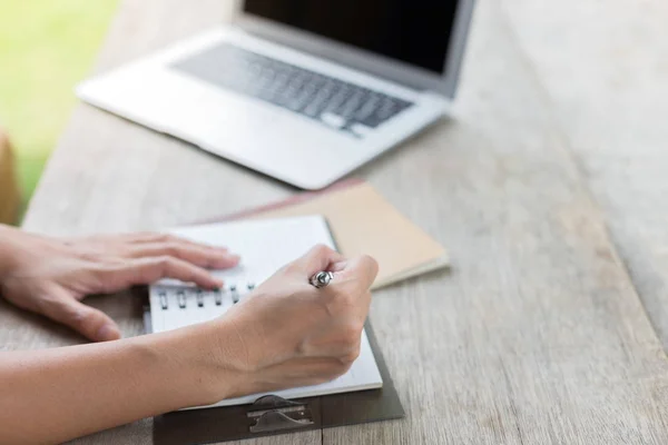 Una mujer sosteniendo un bolígrafo está escribiendo en un cuaderno con una computadora portátil y — Foto de Stock