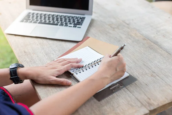 Una mujer sosteniendo un bolígrafo está escribiendo en un cuaderno con una computadora portátil y — Foto de Stock