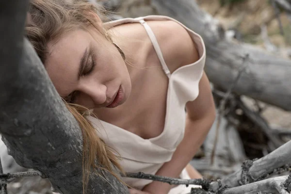 A girl dressed in a beige dress sat on the trunk of a fallen dry tree and leaned against a dry branch. The dry bed of a mountain river. The nature of Altai land. Portrait of a girl with closed eyes.