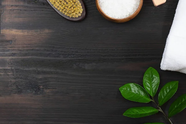 Kadın vücudu ve cilt bakımı, banyo ve spa ürünleri, kaptaki deniz tuzu, beyaz havlu, yeşil yapraklar, siyah ahşap arka plandaki kuru masaj fırçası. Güzellik, kozmetik. Kopyalama alanı ile yatay