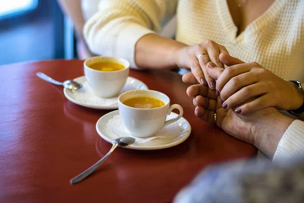 Hombre y mujer tomados de las manos, cerca de los brazos de la pareja en la mesa de madera roja en el restaurante con dos tazas blancas con té en él. fecha en la cafetería — Foto de Stock