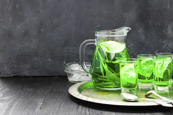 Summer cold green drink with tarragon, mint and lemon in glass decanter and small shots with ice, one in focus in focus. Sparkling beverage, alcohol drink on dark background. Horizontal, copy space — ストック写真