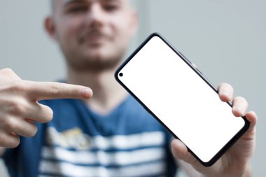 Akıllı telefonunu tutan genç bir adam beyaz ekrana işaret parmağını gösteriyor. Bulanık arka plan odaklı ön kamerası olan akıllı telefon boş ekran. Uygulama ve web sitesi tasarımı için model.