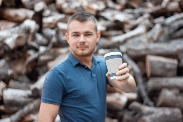 Genç adam arka plandaki testere kütüklerinin üzerine kahvesini koyup elini uzatıyor. Odaklı plastik bardak arka plan bulanıklaşmış.